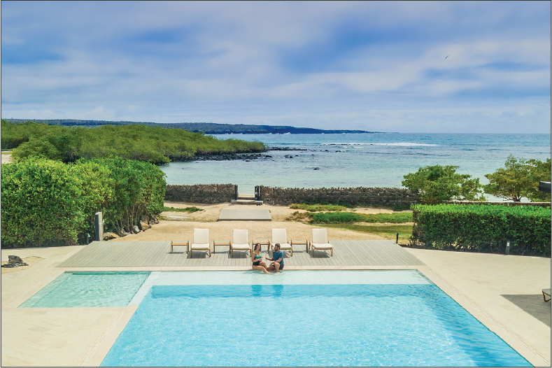 A couple relaxes by the side of the pool at the Finch Bay Hotel. A spectacular view of the ocean fills the background.